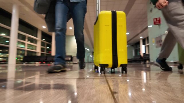 Low angle shot of people with luggage walking through an airport terminal, following a yellow suitcase and showing passengers' legs and feet as they rush to their gate
