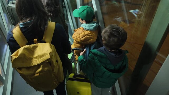 Family of tourists with luggage walking through the airport jet bridge before departure, a mother with her two children embarking on a plane for their vacation trip