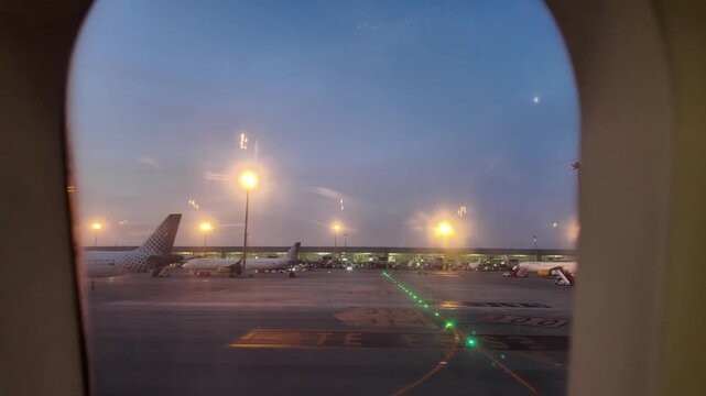 Airplane taxiing on the airport tarmac at dusk, pov from the window with views of the terminal, parked planes and glowing lights before departure or after arrival