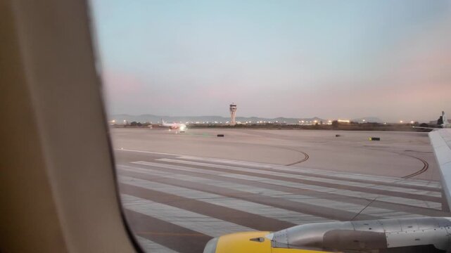 Airplane window view of the runway during takeoff with the air traffic control tower and a scenic pastel sunset sky visible in the distance, showing travel and transportation