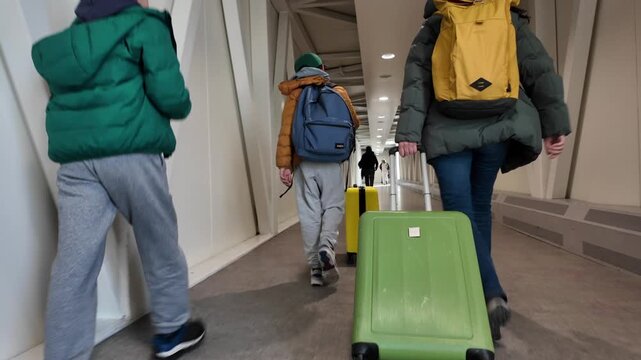 Family with suitcases and luggage walking along the airport jet bridge corridor, a mother and two sons boarding a plane for their vacation journey and trip