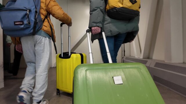 Anonymous travelers pulling wheeled suitcases and carrying backpacks down an airport jet bridge toward the boarding gate, captured from behind in a busy terminal corridor