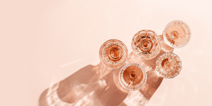 Wine Glasses of rose wine on pink background, sun flare and reflection from sunlight. Aesthetic banner from various crystal glasses, sparkling alcohol drinks, champagne, Summer romantic setting