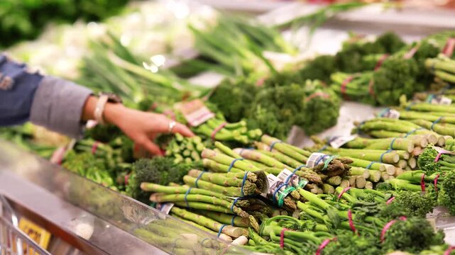 Woman hand picking fresh green broccolini and asparagus at grocery store vegetable aisle.