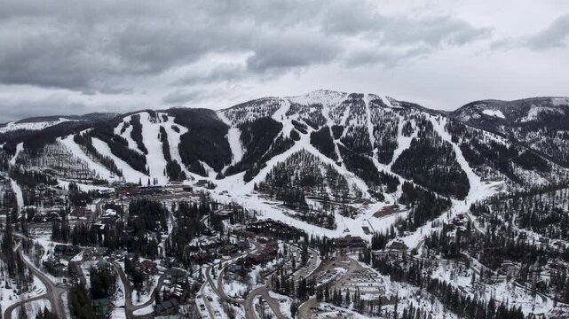 Aerial drone footage captures a mountain ski resort near Whitefish, Montana. Snow-covered slopes with winding ski runs descend into a village of lodges and roads, framed by dramatic clouds.