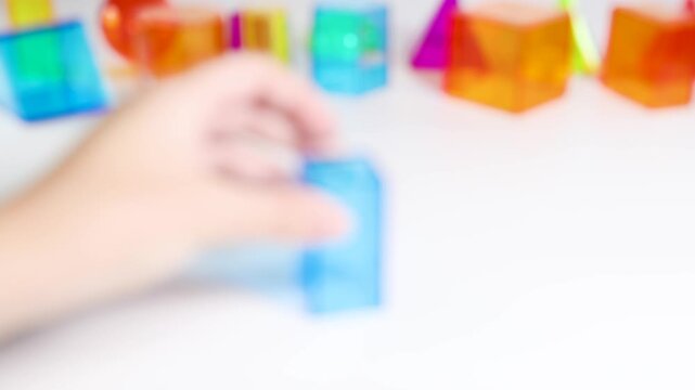 Hand picks up transparent blue geometric block from white surface with other blocks blurred in background.