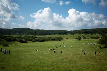Fototapeta premium Lush green field under bright blue sky with scattered clouds. Small group of people relaxing in open grassy area. Distant hills covered in dense green trees and shrubs
