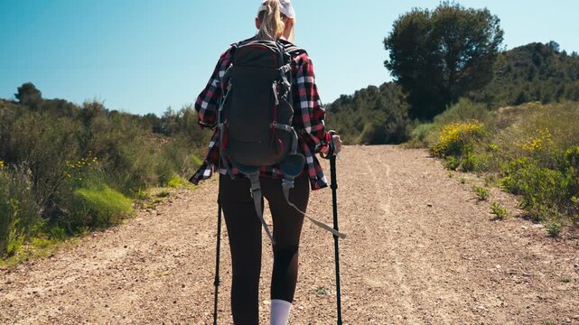 Young woman with trekking poles and backpack walking away from camera on dirt road during sunny day. Concept of outdoor adventure, travel, freedom and active healthy lifestyle in nature