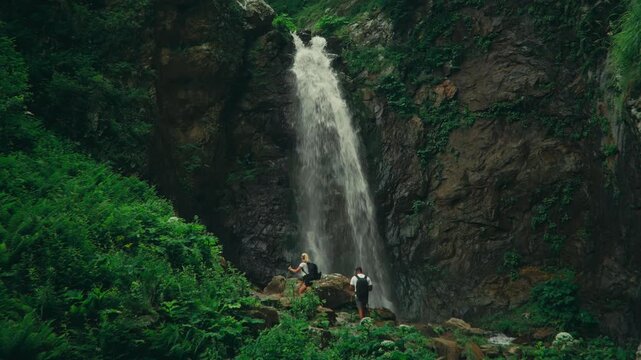 Hikers arrive at a stunning waterfall gorge and stand on an elevated boulder in front of the falls, highlighting scale, power, and immersion in wild natural landscape. Gveleti big waterfall Georgia