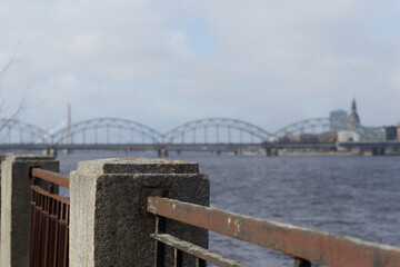 Fototapeta premium Close-up of concrete embankment post and rusty railing beside river, with softly blurred arched bridge and skyline in background.