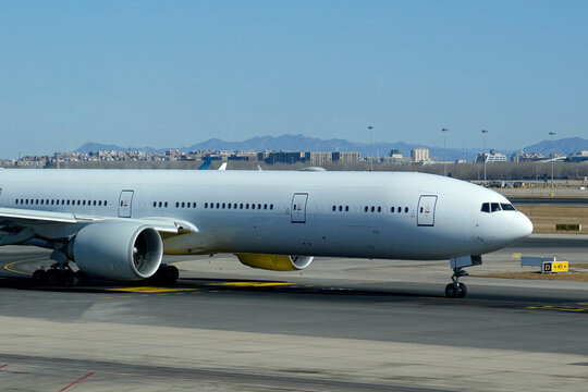 This magnificent passenger jet idles on the sunlit airport ground. Urban architecture and serene mountains unfold behind it, under an expansive blue heaven