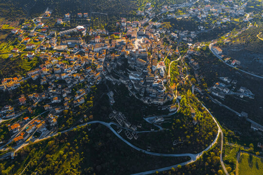 Aerial view of the historic stone buildings and the Piccolomini Castle nestled on a lush green hillside during golden hour in Capestrano, Abruzzo, Italy.