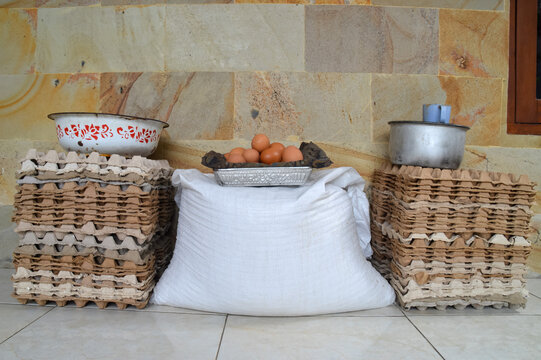 A front view of several fresh eggs gathered in a bowl, resting on top of stacked bags of layer chicken feed with egg crates and farming supplies on a house porch.