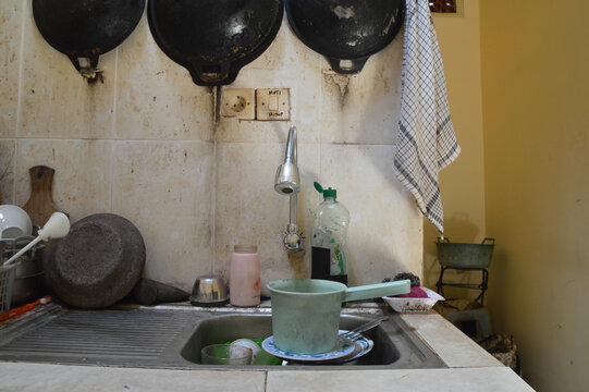 A front eye level view of a chrome flexible swan neck tap positioned above a kitchen sink filled with dirty plates, spoons, and a plastic water dipper ready for washing.