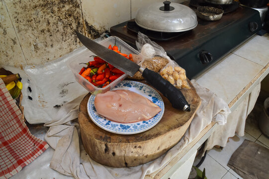 A quarter-left elevated perspective featuring chopped raw chicken, red peppers, and various herbs arranged on a rustic wooden tray alongside a sharp traditional cleaver.