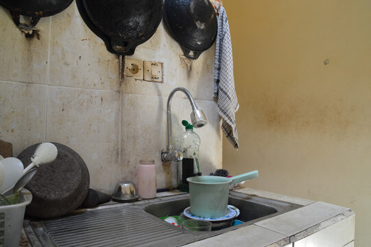 A quarter left perspective showing a flexible goose neck faucet and a stack of used kitchenware including plates and a water scoop in a domestic wash basin area.