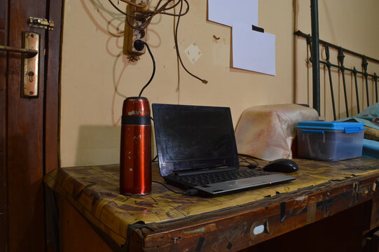 A quarter left angle shot of a modern laptop paired with an external mouse and a mini thermos, neatly arranged on a wooden desk surface within a quiet bedroom environment.