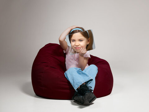 Little girl smiling and holding portrait frame while sitting on bean bag isolated studio background. Concept of identity exploration, self perception, emotions and playful digital communication.