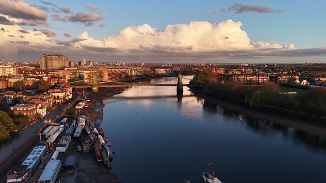 London, UK - April 20 2026: 4K aerial drone shot of the historic Hammersmith Bridge over the River Thames during a vibrant golden hour sunset. Cinematic view of rowing boats on the water, residential 