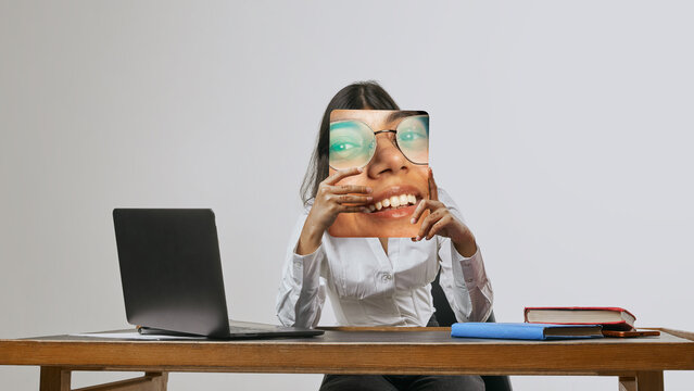 Young woman sitting at desk holding printed smiling face panel office lifestyle portrait. Concept of workplace emotions, professional image, mood masking, communication and corporate identity.