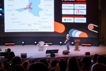 Conference presentation on stage with speaker, large screen graphics, attentive audience in dim auditorium. © Anton Gvozdikov