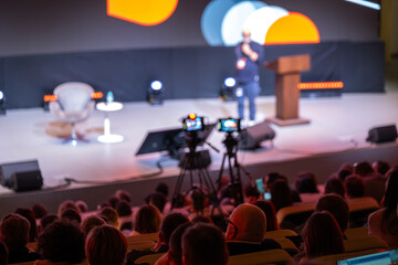 Conference speaker on stage, cameras recording presentation, live event atmosphere, attentive audience, modern lighting and audiovisual production. © Anton Gvozdikov