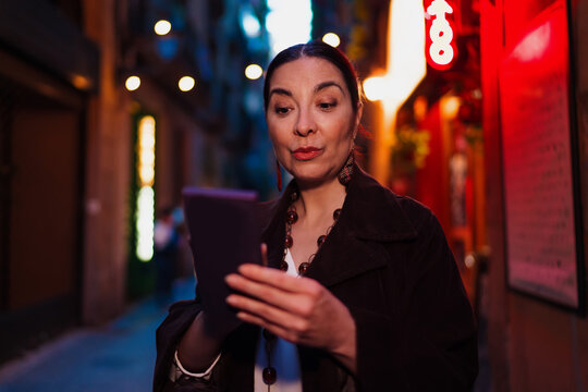 Hispanic woman with intrigued expression reading from tablet on street at dusk, symbolizing discovery, tech fluency and curiosity in modern connected lifestyle.