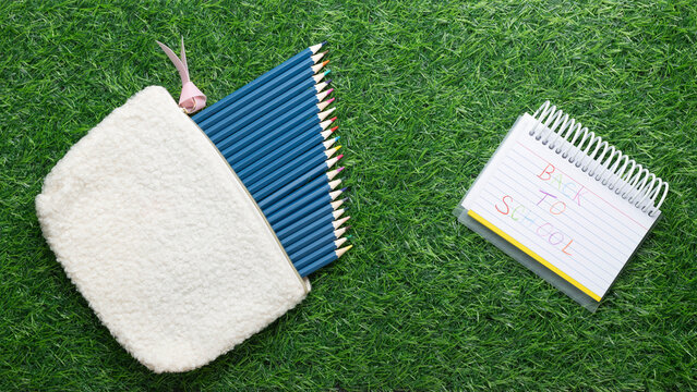 Back to school message handwritten on lined paper.  Multicolored pencils in a cream-colored fabric bag or pouch on green grass.
