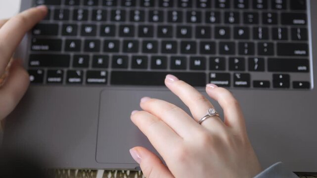 Close-up of hands typing on laptop keyboard. Left hand holds spacebar. Right hand moves over touchpad. Ring visible on right ring finger. Fingernails neatly manicured. Laptop surface sleek and modern