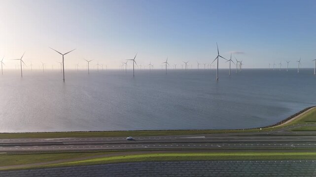Static drone shot from above the IJsselmeer side looking across the dam toward the North Sea in warm morning light.