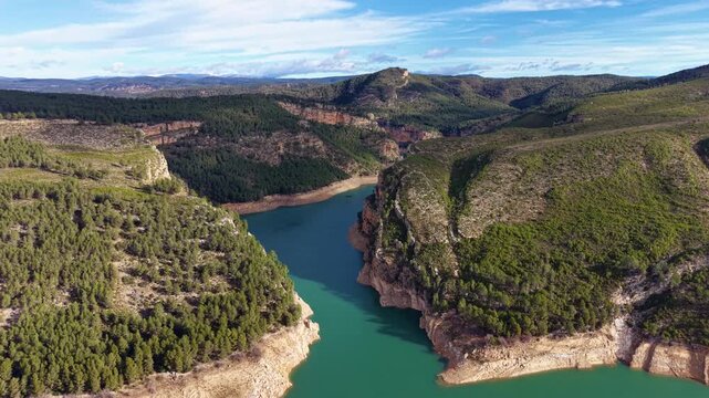 Turquoise reservoir winding through a forested canyon with red rock cliffs and pine-covered hills under a partly cloudy sky in rural Spain