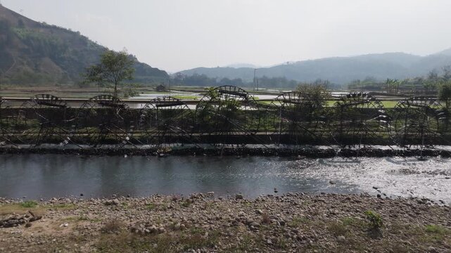 High wide shot of flooded rice fields near Guồng Nước N&agrave; Khương - Bản Bo bordered by dikes and paths, with mountains behind and sunlight glinting off the water.