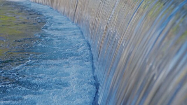 A serene, close-up shot of water flowing over a curved stone weir at Heisei Teien Garden. The water creates smooth curtains and white bubbles, reflecting the soft daylight of a traditional Japanese la