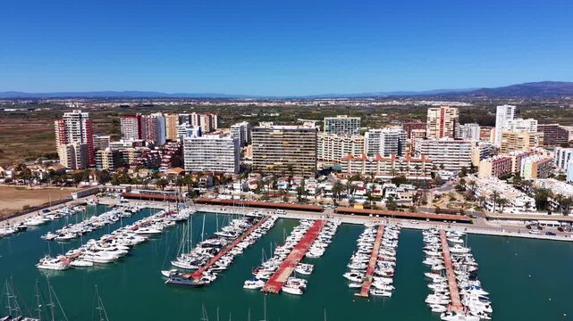 La Pobla de Farnals marina near Valencia, Spain, with rows of sailboats and yachts moored at docks, coastal apartment buildings, palm trees and mountains in the background
