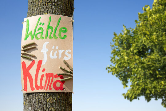 Ein handgemaltes Plakat auf braunem Karton, das mit der Botschaft &bdquo;W&auml;hle f&uuml;rs Klima&ldquo; an einem Baumstamm befestigt ist.