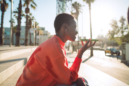 Woman in bright sweater sits on outdoor steps, smiling while holding smartphone horizontally for voice interaction, showing relaxed use of wireless audio tech without visual focus on screen.