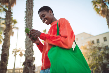 Black woman looks down smiling at her green smartphone while walking, showing immersive mobile media interaction, modern wireless tech use, and everyday digital connection in sunlight. © BullRun