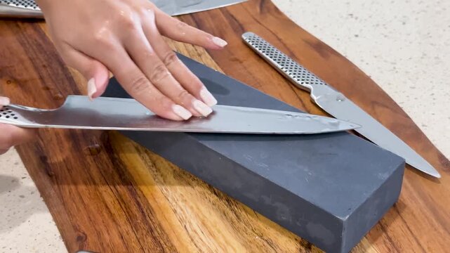 Close up of person hands sharpening chef knife on whetstone block over wooden board.