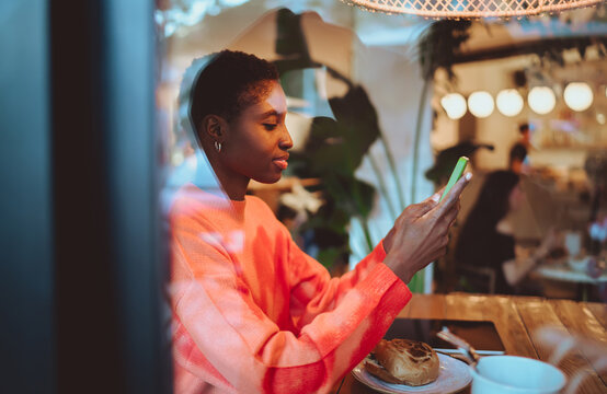 Smiling young woman sitting at a table in a modern cafe with green plants, preparing to enjoy a burger, representing daily city life and authentic dining experiences in a bright atmosphere.