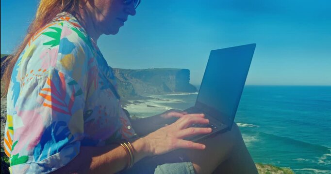 Digital Nomad in Paradise: A digital nomad enjoys remote work, typing away on a laptop while perched atop a scenic cliff, overlooking a breathtaking coastal panorama of sea and sky.