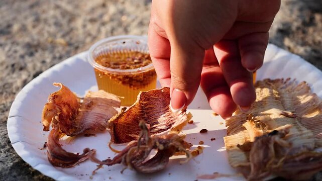A close-up shot of a person picking up dried squid with their fingers from a white plate, showcasing street food culture with delicious sauces.