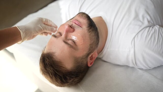 Cosmetologist applying eye patch on one eye of male patient. Close-up video of skincare procedure in aesthetic clinic, focusing on hydration and under-eye treatment.