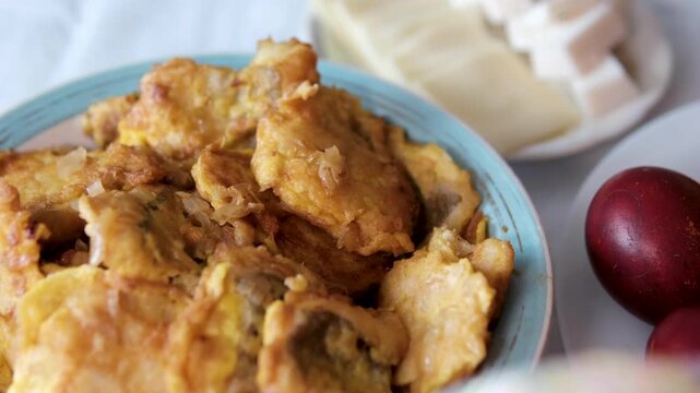 Fried fish or meat in batter is served on a blue plate, with a side plate of cheese and Easter eggs nearby. Close-up of a festive table setting with traditional homemade dishes and holiday atmosphere.