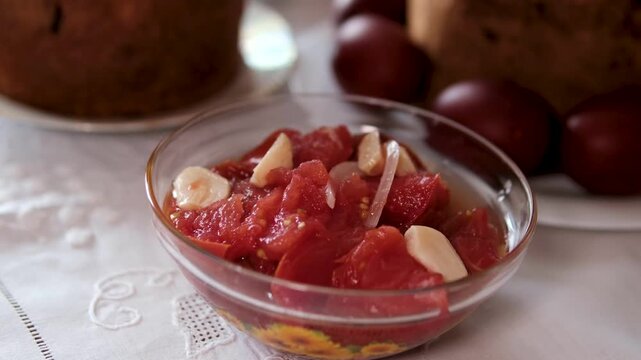 Pickled tomato salad with garlic is served in a glass bowl on a white tablecloth near Easter bread and dyed eggs. Close-up of a traditional homemade dish as part of a festive holiday table.