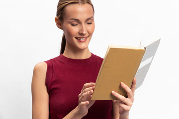 Obraz premium Young woman in red sleeveless top reading a book with a smile on white background. Concept of education, leisure, knowledge, happiness, and learning in casual lifestyle.