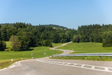 Naklejka premium Winding rural road stretches green fields forest clear blue sky create scenic perspective sense freedom. Peaceful countryside landscape gentle hills asphalt invites travel exploration calm journey