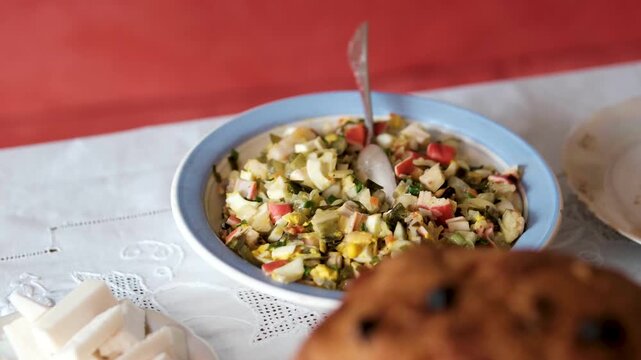 Elderly female hands place a seafood salad with crab sticks, eggs, and mayonnaise onto a festive Easter table near traditional bread. Close-up of a homemade dish added to a white tablecloth setting.
