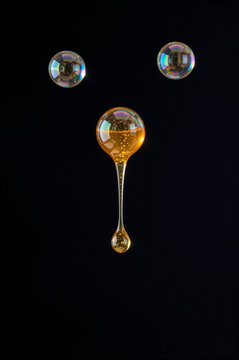 Amber viscous liquid forms an elongated, gravity-defying drip from a suspended golden droplet against a void of deep black. Prismatic specular highlights dance across the translucent surface.