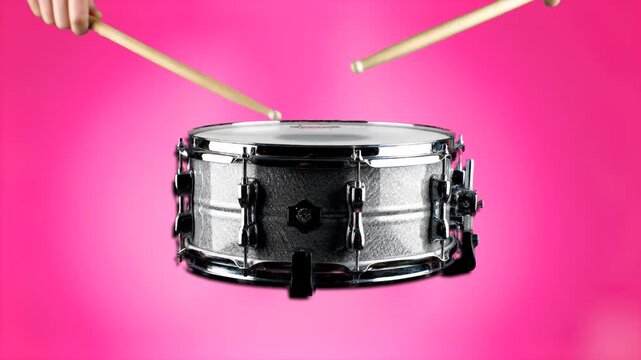 A close-up shot of a metallic snare drum being struck by drumsticks against a solid pink background. This vibrant studio shot is perfect for musical themes, lifestyle content, or modern artistic motio