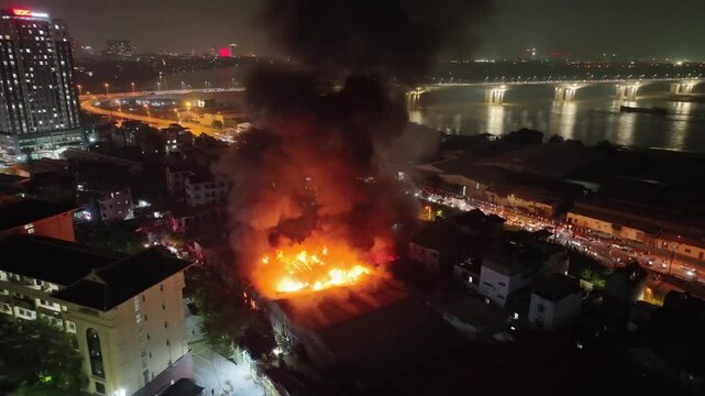 Massive nighttime fire near riverfront in Hanoi, bright flames illuminating nearby buildings and casting reflections across water and skyline.
